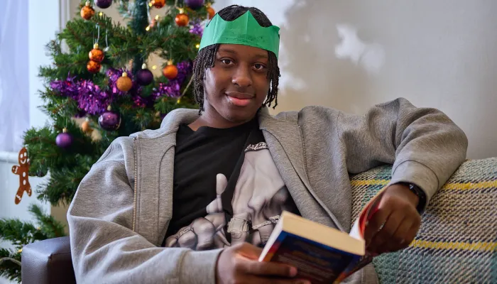A young person sits reading a book they received for Christmas on their sofa next to the Christmas tree. They're still wearing the paper hat from their cracker, and smiling at the camera.