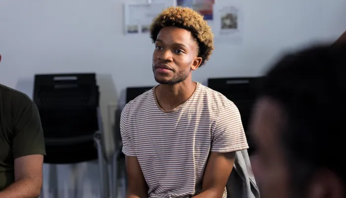 Person seated in a group discussion, wearing a striped T-shirt and a smartwatch, with other participants partially visible in the foreground and background of a meeting room.