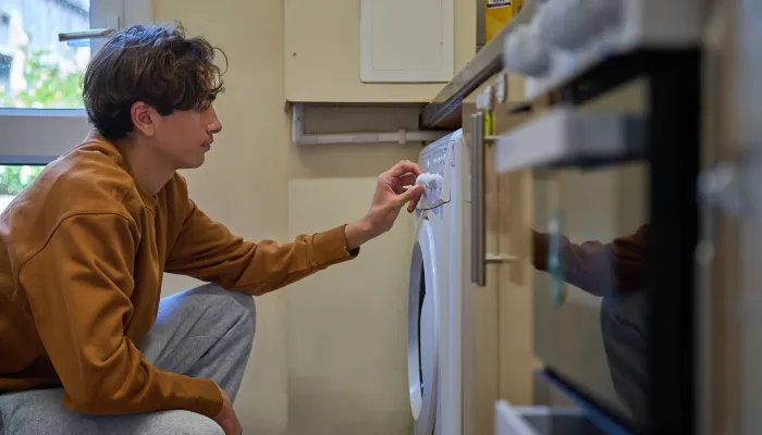 A young person kneels by the washing machine and sets the laundry program