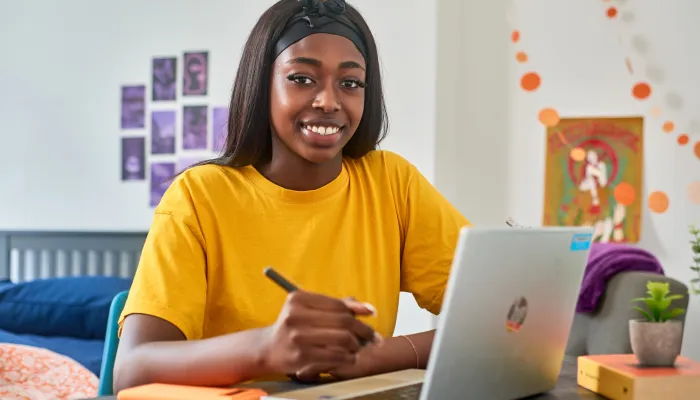 A young person sits at a desk with a notepad and pen and a laptop for studying