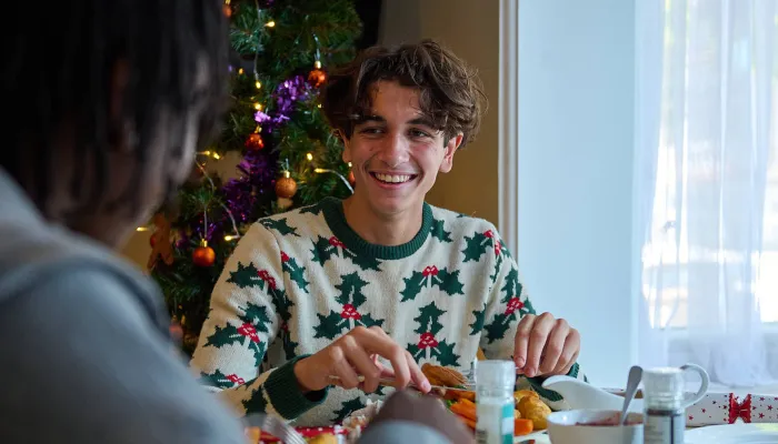 A young man sits at a table eating Christmas dinner, smiling at another person across from him. There is a Christmas tree behind him and he is wearing a Christmas jumper with holly on it. 