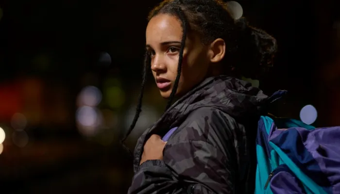 A young woman stands outside at night, wrapped in warm winter clothing and carrying a bag.
