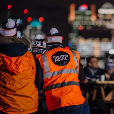 Two people stood with their backs to the camera wearing high vis jackets and bobble hats with London's skyline in the background.