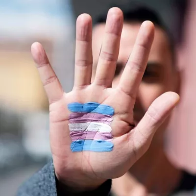 Photograph shows a person extending their hand towards the camera with a trans flag painted on their palm.