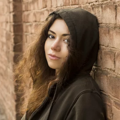Young person with long brown curly hair in a brown hoodie standing against a brick wall