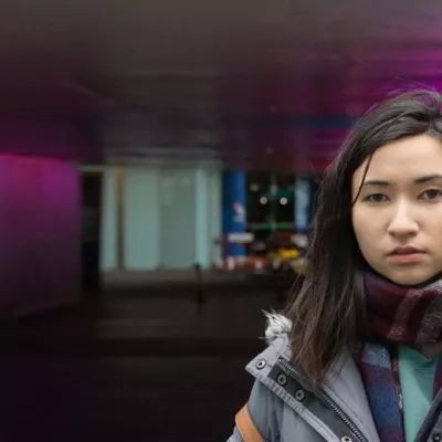 Homeless young person standing underneath an underpass, wearing a scarf and looking to the camera.