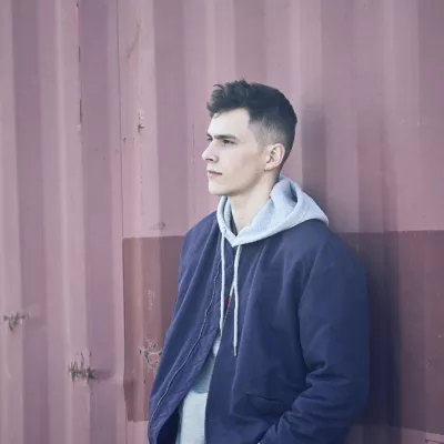Young person in blue jacket and grey hoodie stands against a corrugated metal wall