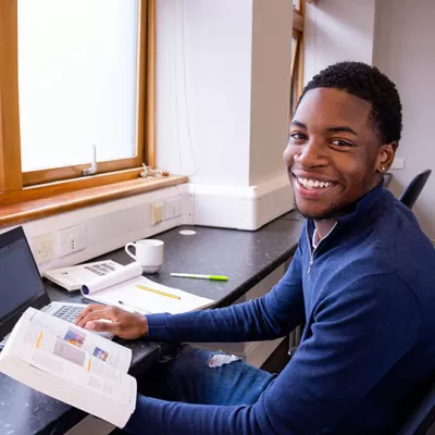 A young person sits at a desk with a laptop and notebooks, studying for their lessons