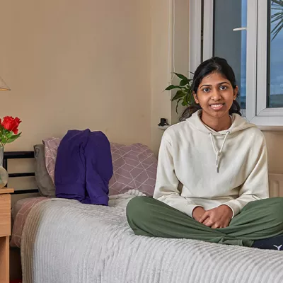 A young person sits on a single bed in their cosy Centrepoint room, smiling at the camera