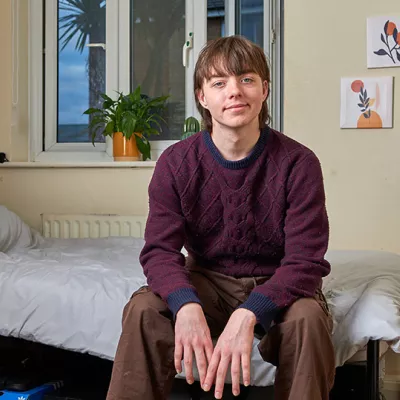 A young person sits, smiling, on a single bed in their Centrepoint room. There are plants on the windowsill and small pictures on the wall to give a cosy feel.