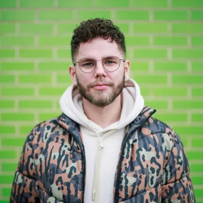 A portrait of Radio 1’s Dean McCullough directly facing the camera in front of a green brick wall