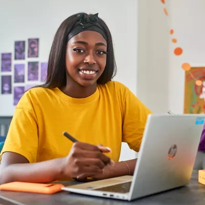 A young person sits at a desk with a laptop, pen and notepad in their own room