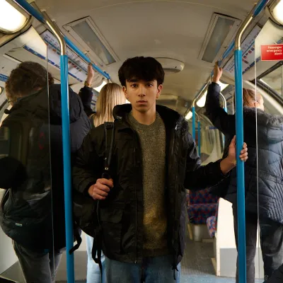 A young homeless person stands looking scared in a busy tube train carriage holding a bag of their belongings