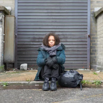A homeless young women is sat on a curb with her rucksack next to her. She looks tired and is looking straight on.  