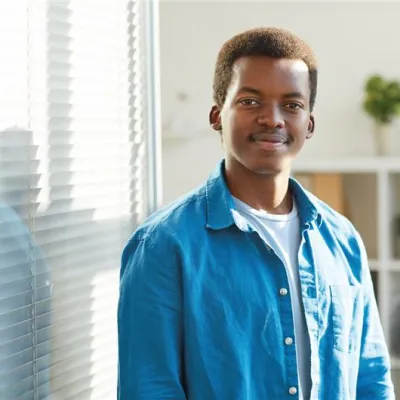 A young man standing next to a window with blinds, wearing a blue button-up shirt over a white t-shirt. He is smiling softly and looking at the camera, with a bookshelf and a potted plant in the background.
