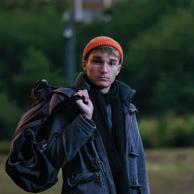 A young person standing outdoors on a patch of grass, carrying a duffel bag over one shoulder and holding a rolled-up sleeping bag. They’re wearing a grey parka, green trousers, a scarf, and an orange beanie, with trees in the background.