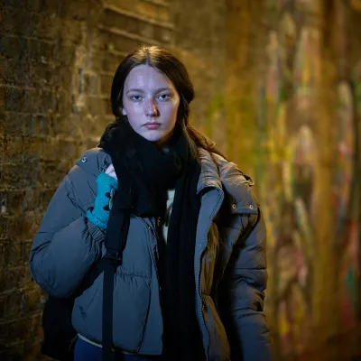 A young woman, NB, stands in a dimly lit alley with a brick wall covered in graffiti. She wears a thick grey puffer jacket, black scarf, and teal fingerless gloves, looking directly at the camera with a serious expression.