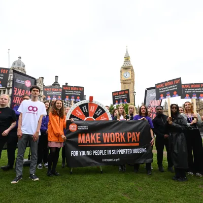A group of people standing outside the Houses of Parliament holding a banner that says 'Make Work Pay for young people in supported housing' and placards