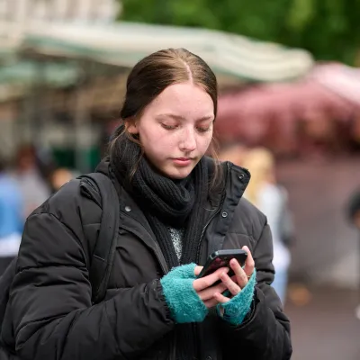 A girl dressed in a thick coat and frayed blue gloves looks down at her phone. She carries a backpack, and is alone with a busy street behind her.