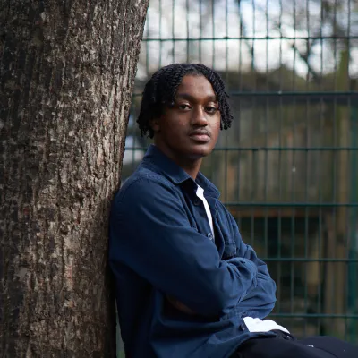 A young person sits in a park, leaning against a tree