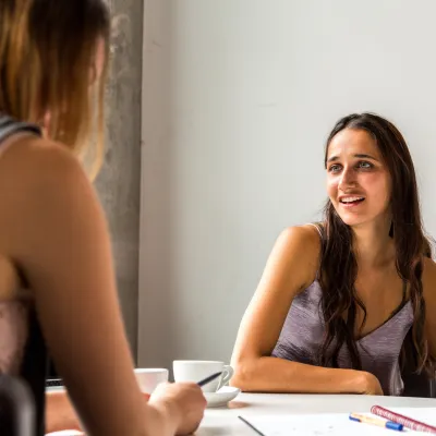 Two people sit across from each other at a white table in a bright indoor setting. One person is in the foreground, partially out of focus, while the other is seated facing the camera. White coffee cups, a notebook, and a pen are on the table, suggesting a casual conversation or meeting. Natural light illuminates the scene against a plain light-colored wall.