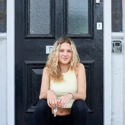 A young woman sits on the doorstep outside her home, holding house keys and looking peaceful
