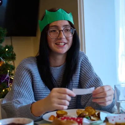 A young person reads a joke from a Christmas cracker over dinner, with a sparkling Christmas tree in the background