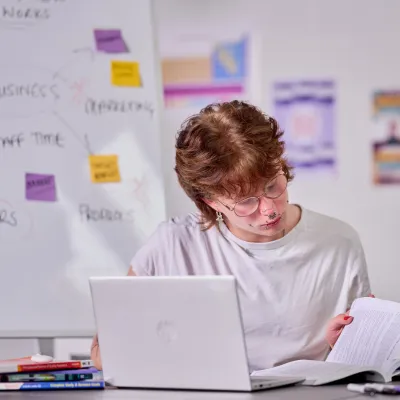 A young person studies with a book and laptop at their desk
