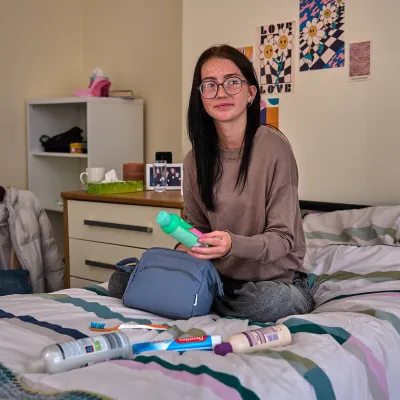 A young person sits on a bed holding personal hygiene items like shampoo