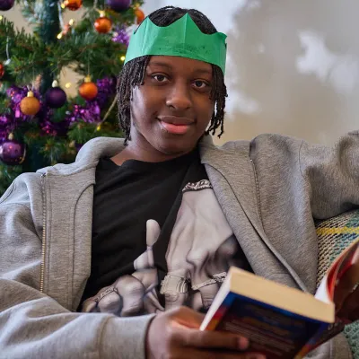 A young person sits reading a book they received for Christmas on their sofa next to the Christmas tree. They're still wearing the paper hat from their cracker, and smiling at the camera.