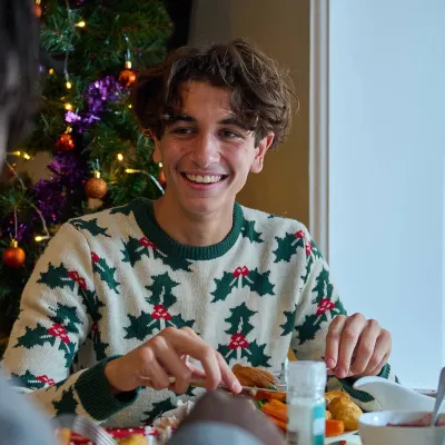 Young people wearing festive jumpers smile while eating their Christmas dinner at the table