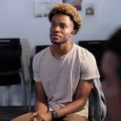 Person seated in a group discussion, wearing a striped T-shirt and a smartwatch, with other participants partially visible in the foreground and background of a meeting room.