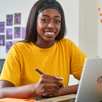 A young person sits at a desk with a notepad and pen and a laptop for studying