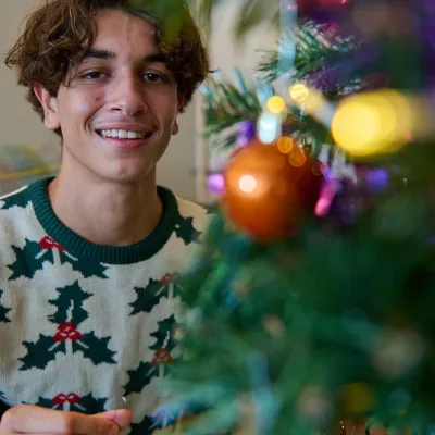 A young man in a Christmas jumper sits in front of a Christmas tree