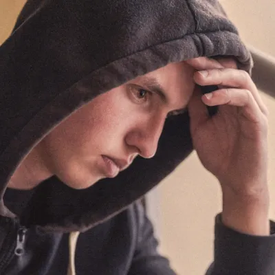 Close-up of a person in a dark hooded sweatshirt sitting near a window, resting one hand against their forehead while looking downward. Natural light from the window illuminates the side of the face, with a blurred indoor background and stair railing visible.